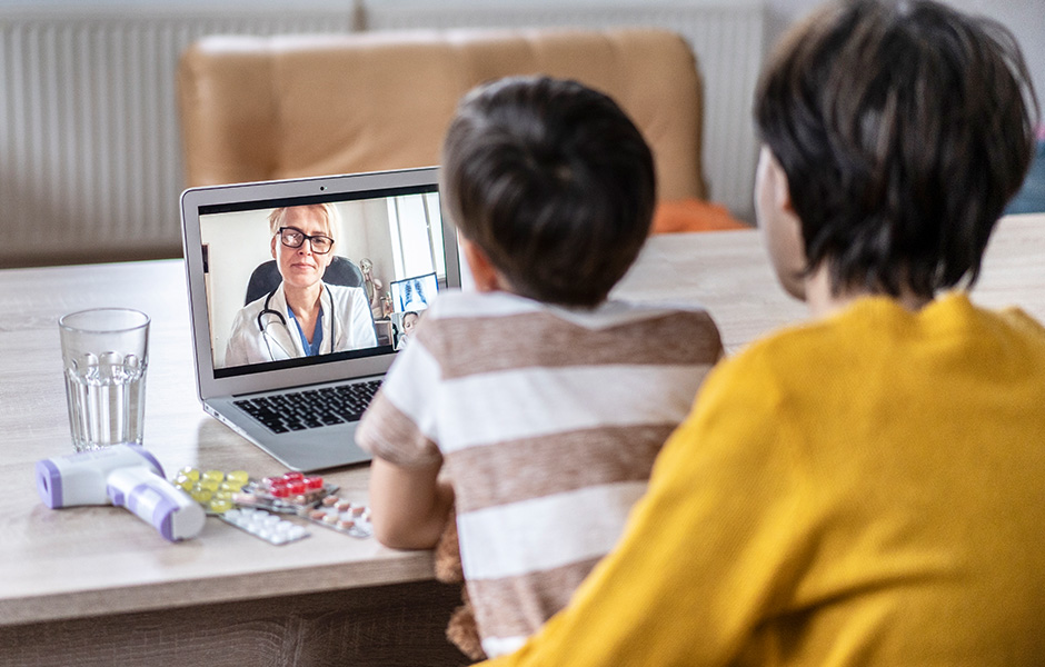 Mother and child at home office with digital thermometer, view a female doctor in lab coat on laptop screen for telehealth visit