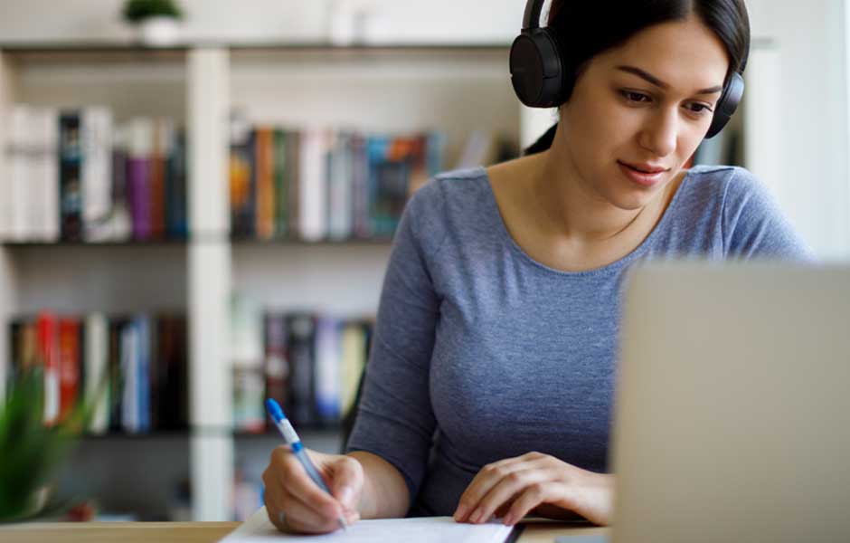 Woman at computer with headphones taking notes.