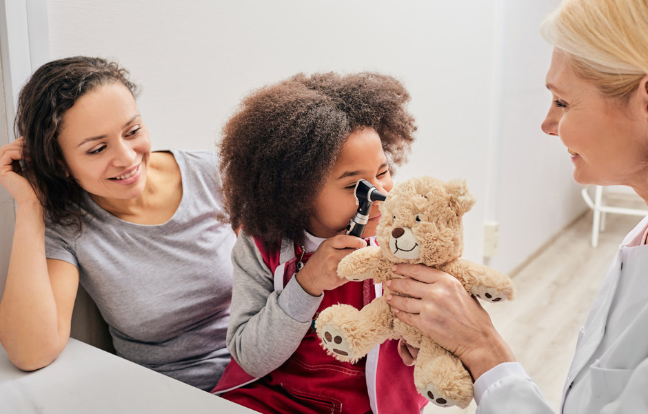 Una médica permite que una pequeña paciente use el otoscopio para examinar a un oso de peluche.