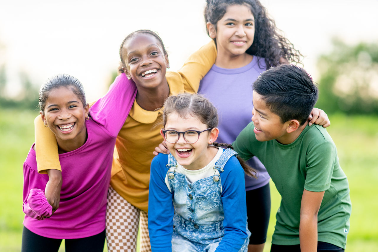 A small group of multi-ethnic school aged children huddle together outside as they pose for a portrait. They are each dressed casually as they laugh and smile in the warm summer air.