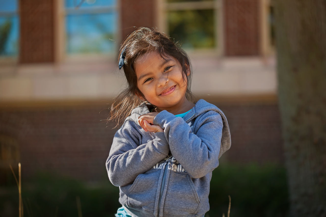 Child wearing a gray hoodie standing outside near a tree with a brick building in the background.