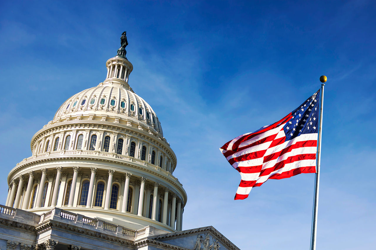 The iconic US Capitol building is shown under a clear blue sky, symbolizing American democracy. An American flag waves prominently in the foreground, adding a patriotic touch to the scene. The architectural details of the Capitol dome are clearly visible, emphasizing its historical significance.
