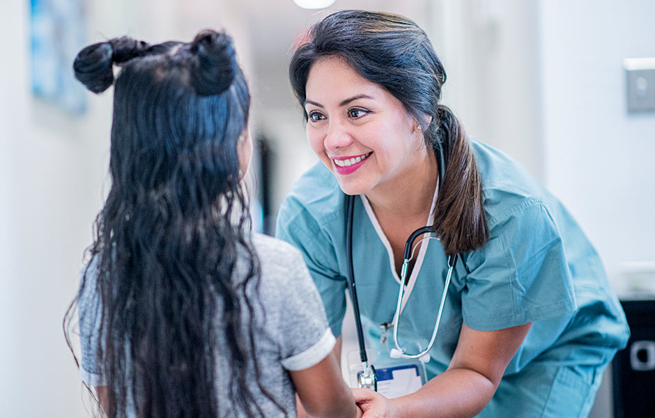 Young female health worker in scrubs leans down to reach eye level and smiles at a female patient. 