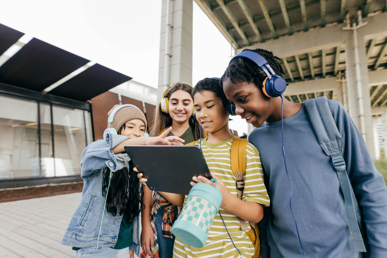 Four children standing outside under an overpass, wearing headphones and sharing a tablet device.