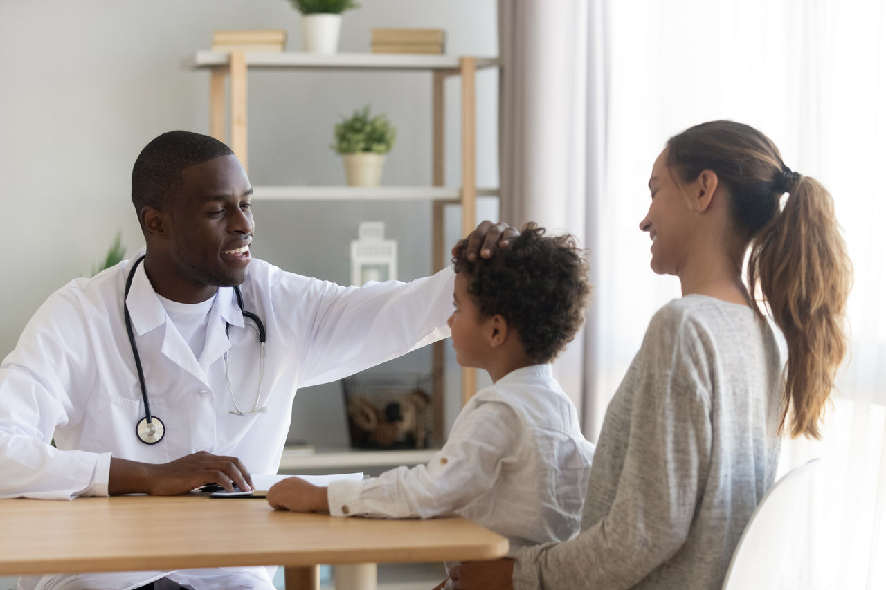 Smiling african american male doctor stroking head of little preschool cute kid patient, satisfied with medical treatment result or boys polite behavior. Happy mom with small son on medical check up.