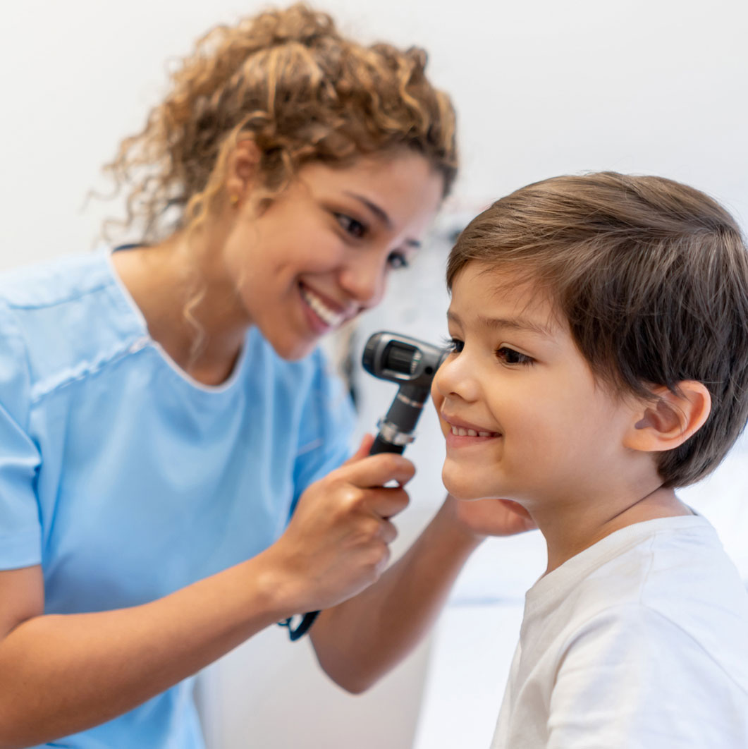 Female health care provider wearing scrubs looks into a young boy's ear with an otoscope.