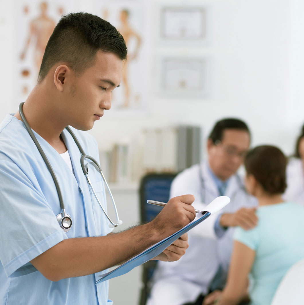 Male nurse in scrubs transcribes while male doctor in lab coat examines patient in the background. 