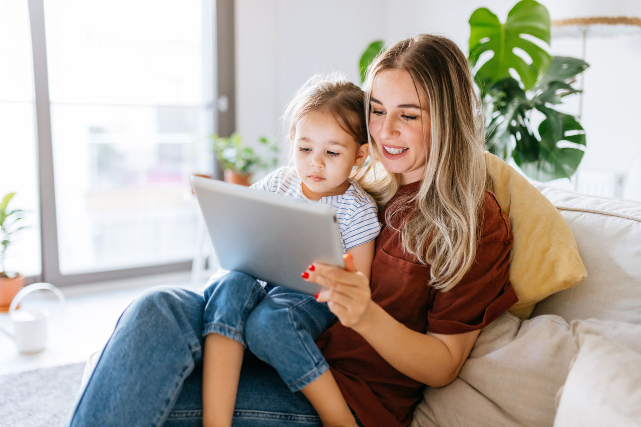 Madre e hija usan una tableta digital juntas. Están sentadas en el sofá en casa. La hija está sentada en el regazo de su madre. Muy feliz y sonriente. Primer plano con recorte ajustado
