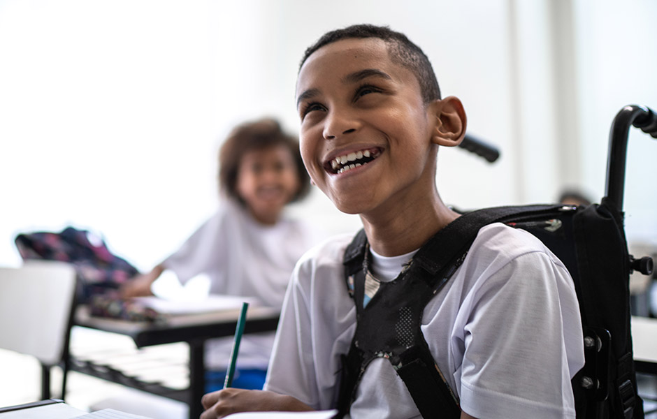 Middle school-aged boy with special needs who uses wheelchair in a classroom, smiling and writing with a pencil