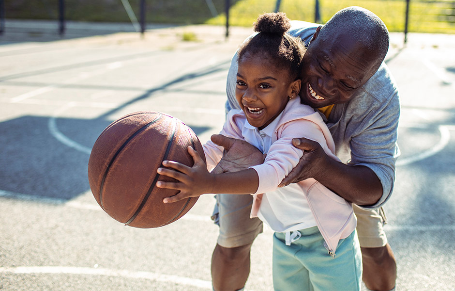 Father helps his young daughter who holds a basketball on a basketball court