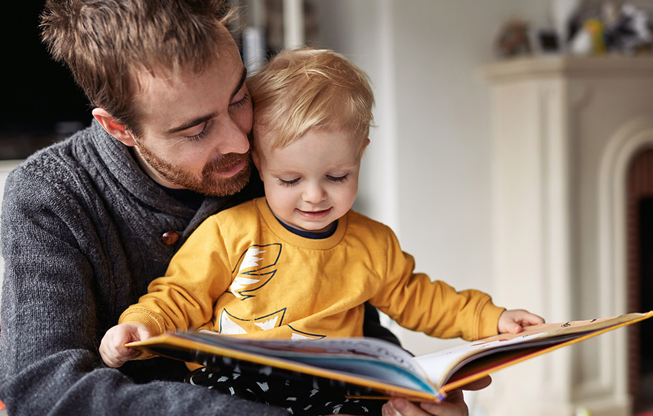 Dad reads to toddler who sits on his lap and holds the book at home before bedtime