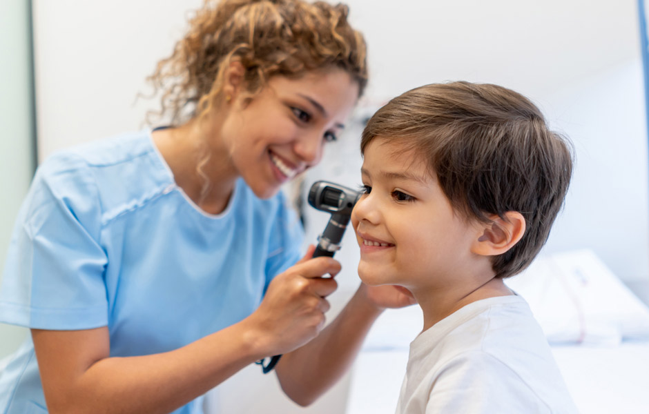 Female health care provider wearing scrubs looks into a young boy's ear with an otoscope.