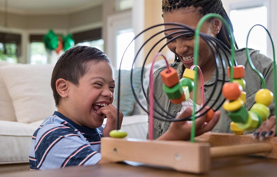 Young boy with special needs sits next to his mom at home playing with a table-top toy and laughing