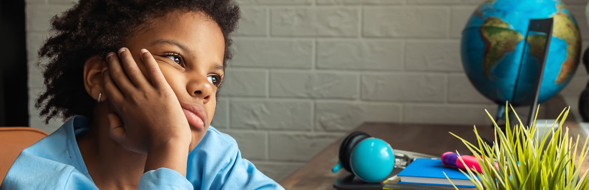 School-aged girl sits alone at desk with chin in hand, headphones set aside, and looking worried.