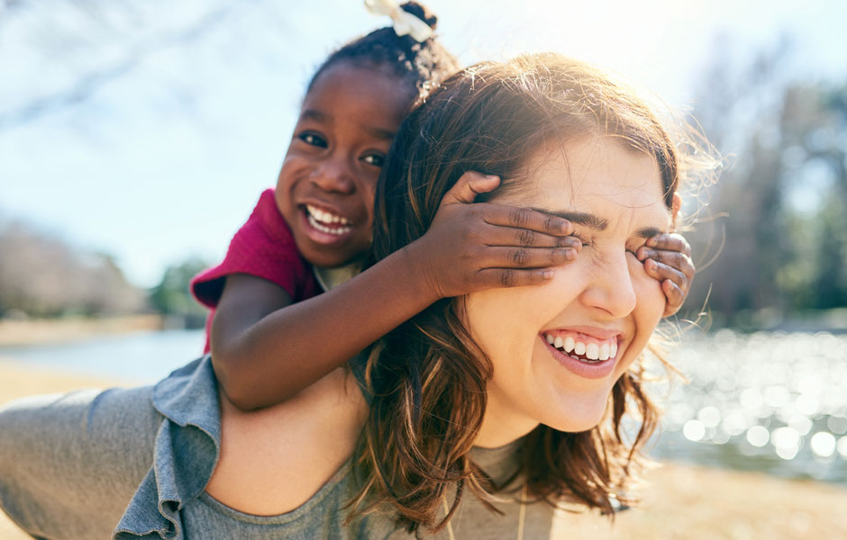 Toddler girl rides piggyback on mom's shoulders and tries to cover mom's eyes. 