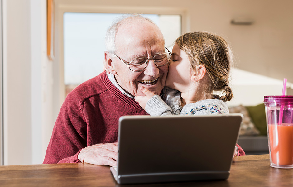 Little girl gives smiling grandfather a big kiss as he looks at laptop
