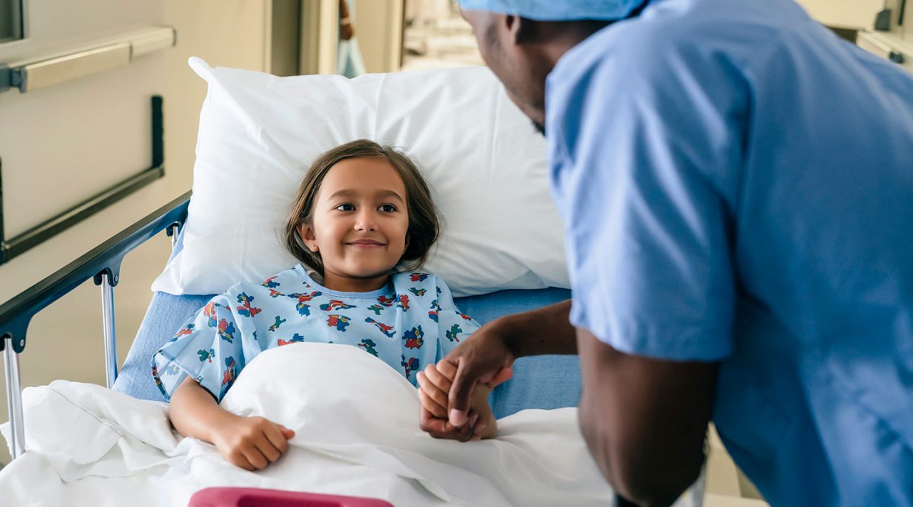 Small girl in a hospital gown and bed talks to healthcare provider who stands at her bedside dressed in scrubs and cap 