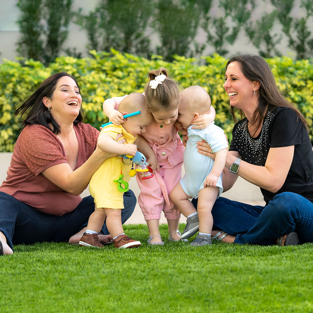 Two moms sit on grass and help three toddlers stand up and hug.