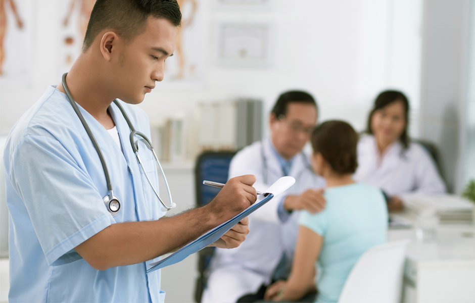 Male nurse in scrubs transcribes while male doctor in lab coat examines patient in the background and a female doctor sits next to them. 