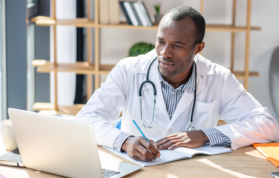 Male doctor looks at laptop and writes in notebook