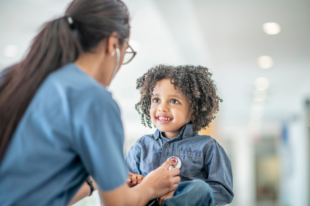 Doctor listening to Pediatric Patients Heart A black toddler boy sits on a table during a medical examination. A female medical professional is using a stethoscope to listen to his heartbeat. The child is smiling up at his doctor.