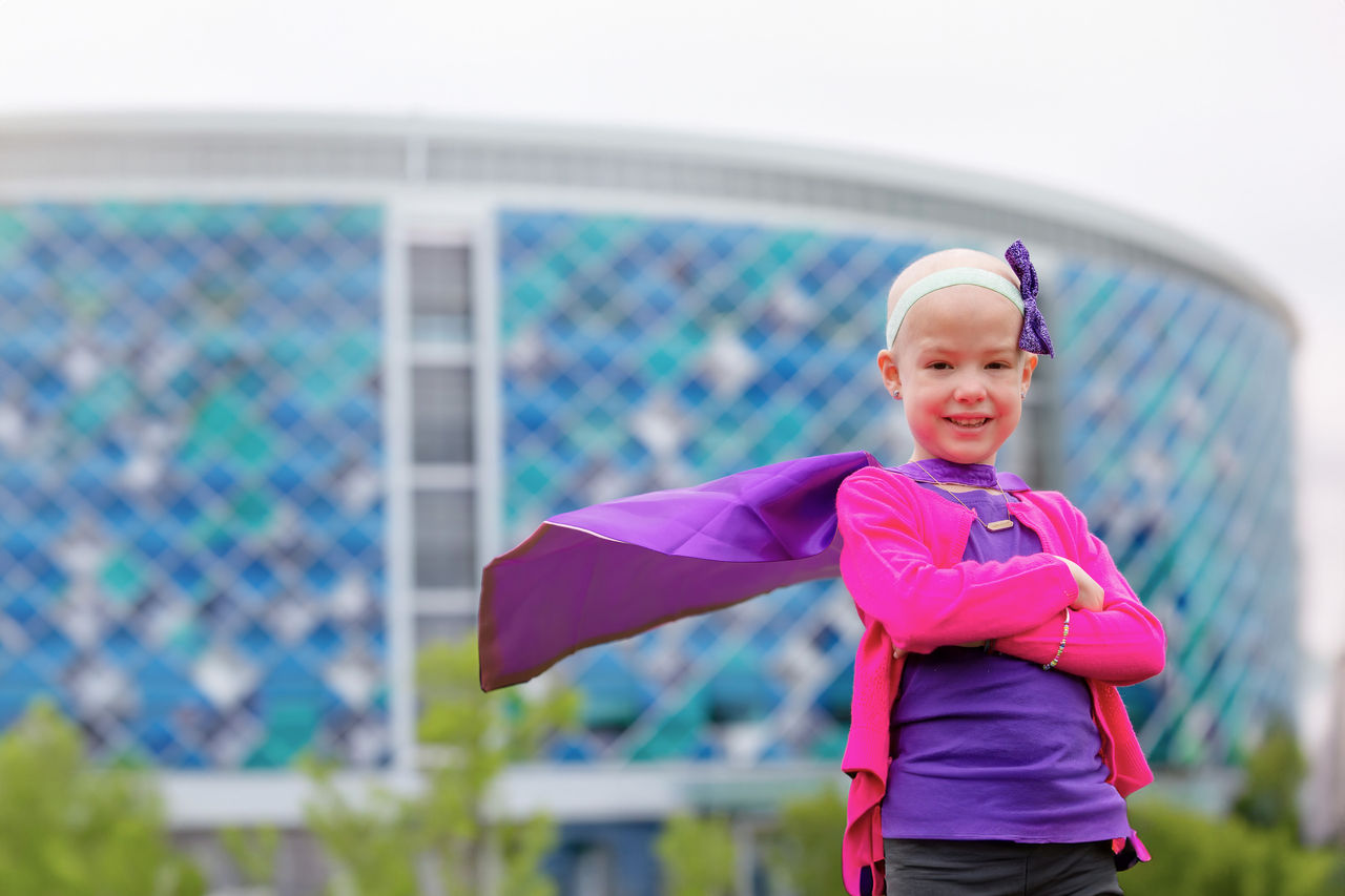 Pediatric cancer patient posing like a superhero with a cape in front of Nemours Children's Hospital Delaware.