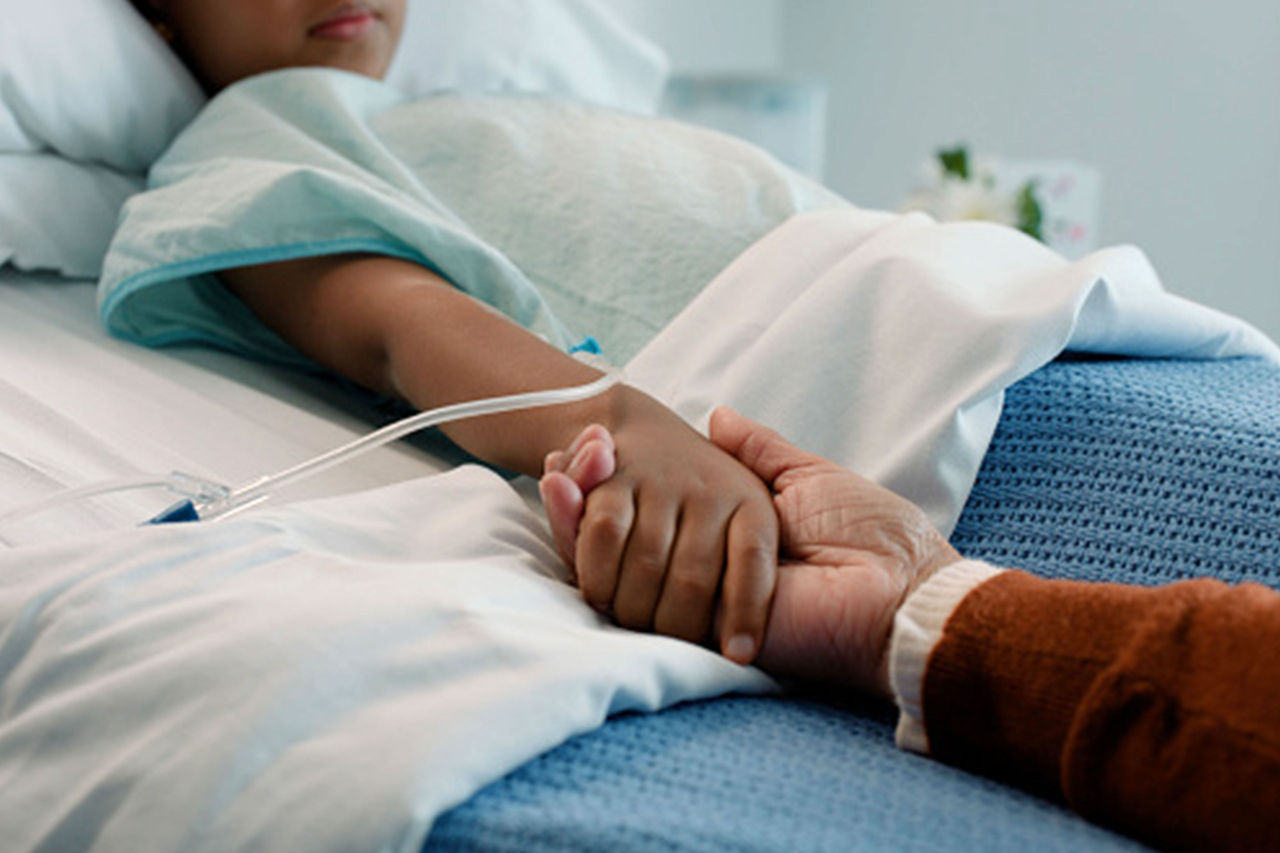 Parent holding childs hand as child lays in hospital bed with an IV in their arm