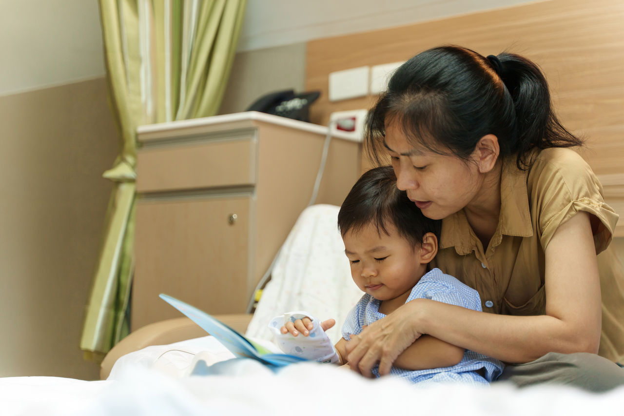 Un adorable niño asiático se enferma por un virus; su madre lo cuida y le lee un libro. Recibió antibióticos y una infusión de solución salina en el hospital, y espera para recuperarse. Concepto médico de atención médica del bebé.