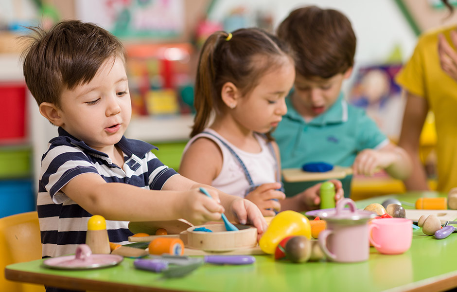 Three preschool-aged kids doing crafts on a classroom table, one boy colors and one girl sculpts with clay