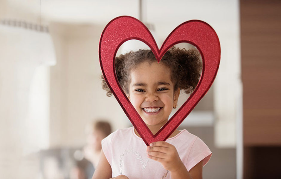 Young girl holds up a paper cut-out of a heart to frame her laughing face.