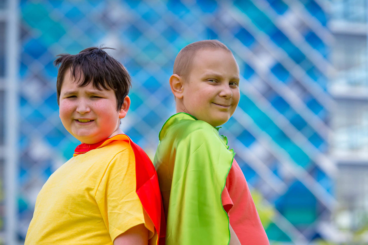 Two pediatric cancer patients standing back-to-back with superhero capes.