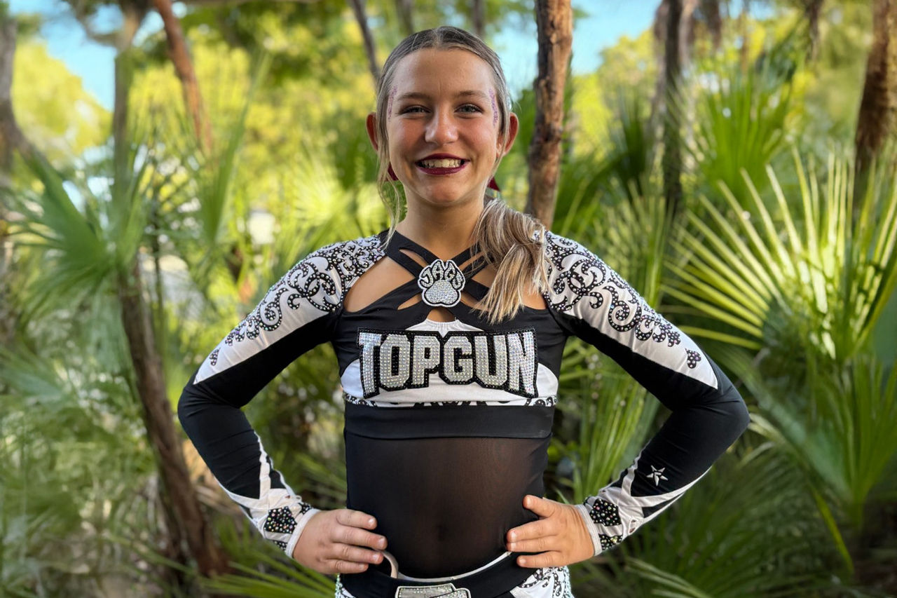 Patient photo of Kaylee wearing a cheerleader uniform standing in front of foliage.