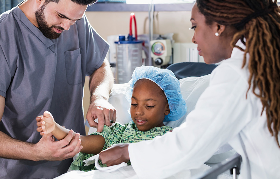 Female surgeon in a lab coat is assisted by a male health worker in scrubs to help a young male patient in a hospital bed, gown and surgical cap get ready for surgery. 