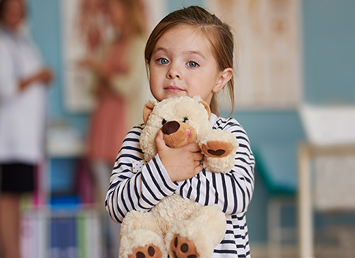 Young girl holds stuffed teddy bear, mom and doctor in white lab coat talk in background
