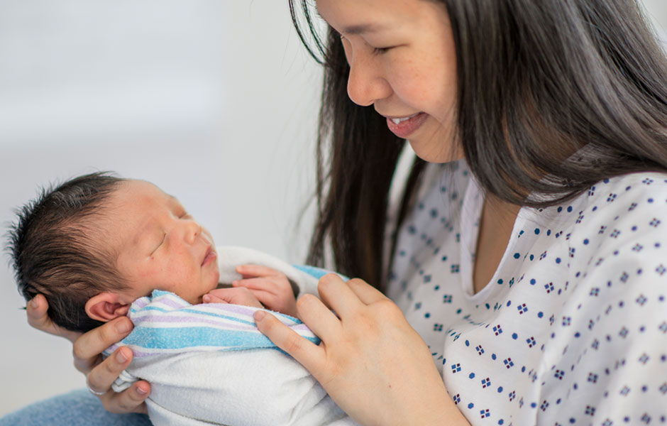Mother in hospital gown smiles at her newborn. Mother in hospital gown smiles at her newborn who is swaddled in a hospital blanket.