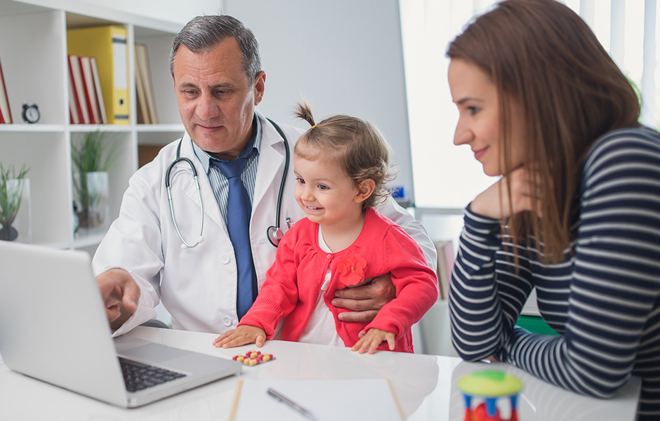Male doctor holds toddler girl as they look at laptop with mother Male doctor holds toddler girl as they look at laptop with mother