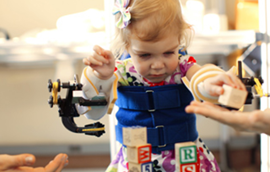 Female toddler wearing body and arm braces, handing a block to rehabilitation therapist