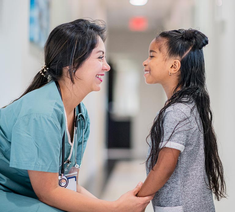 Female doctor in surgical scrubs and stethoscope kneels down and holds the hand of school-aged female patient in hallway. 