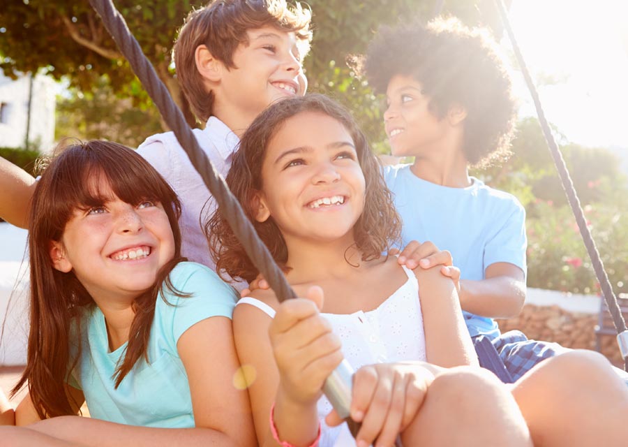 Four school-aged children of different genders and ethnicities sitting on a tire swing together in a sunny  park.