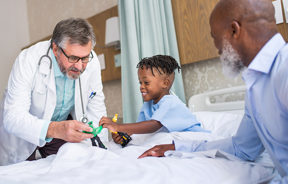 Preschool-aged male patient in hospital bed looks at equipment demonstrated by male doctor while grandfather looks on smiling