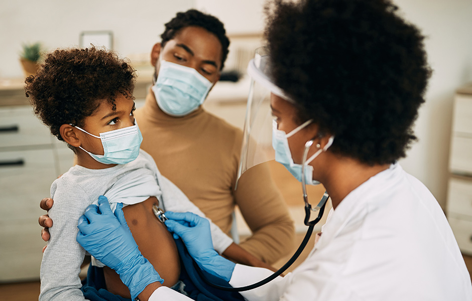 Female doctor examines a toddler while dad holds the baby, all wear face masks