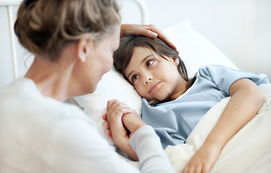 School-aged child in hospital bed holds hands with mother at bedside. School-aged child in hospital bed and gown looks at mother who sits at bedside holding the child's hand.