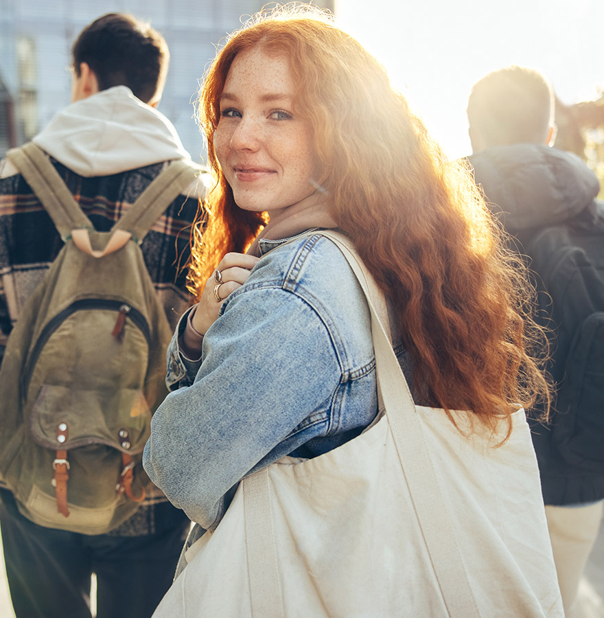 Teenaged girl carrying tote, is stopped to smile for camera while walking behind male classmates who are wearing backpacks. 