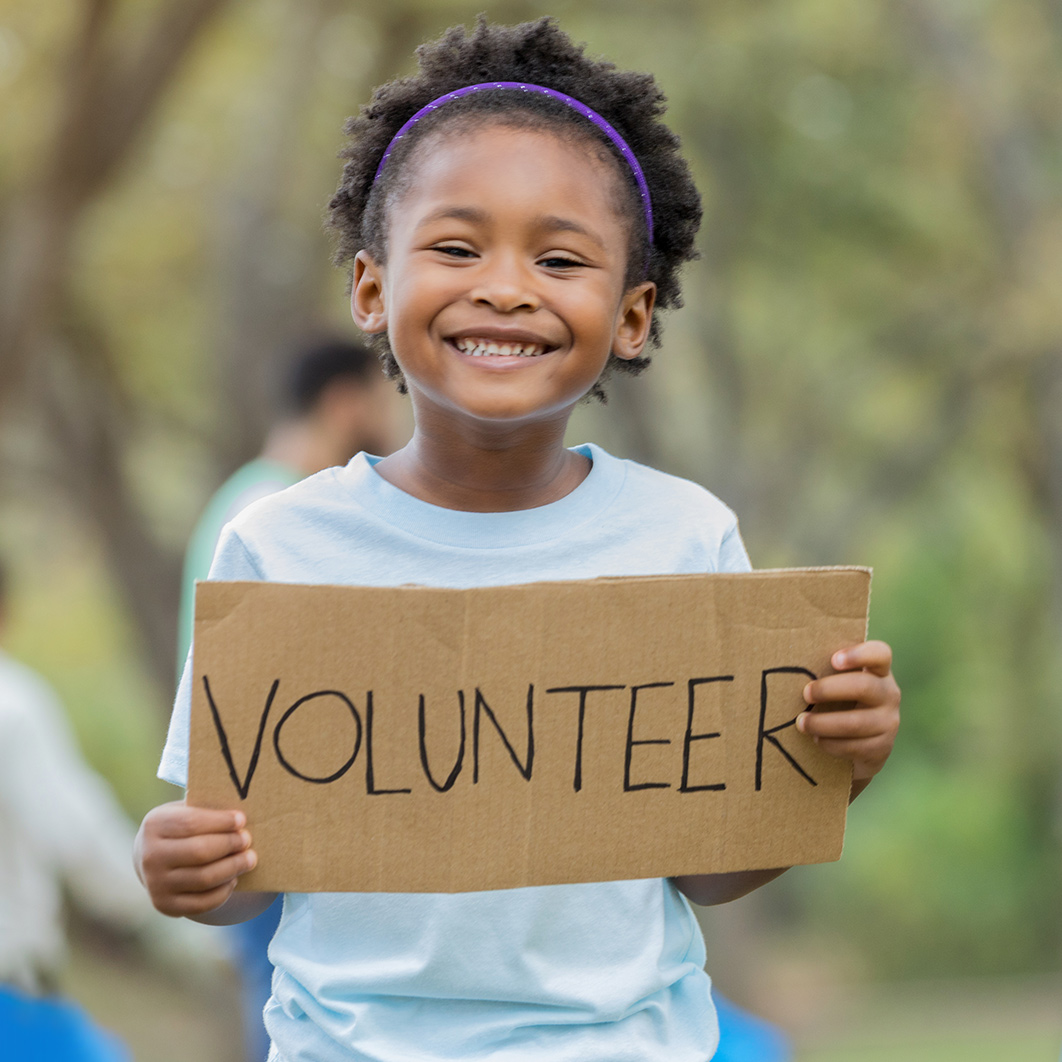 Proud school-aged girl holds up a handmade cardboard sign that reads, "volunteer" 