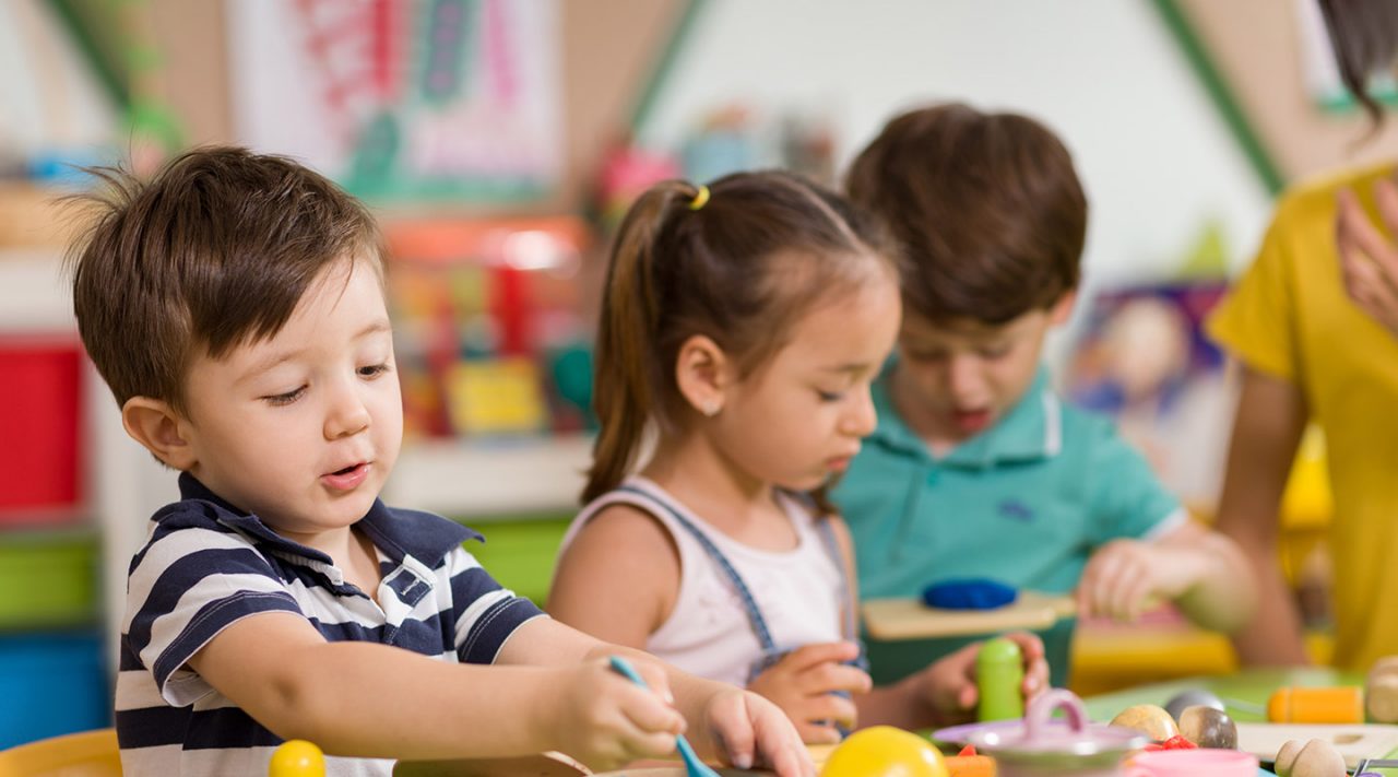 Three preschool-aged kids doing crafts on a classroom table, one boy colors and one girl sculpts with clay